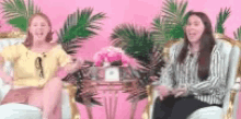 two women are sitting in chairs in front of a pink background with palm trees .
