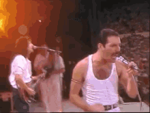 a man in a white tank top is singing into a microphone on a stage with other people playing instruments .