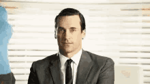 a man in a suit and tie is sitting in front of a window with blinds .