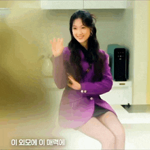 a woman in a purple jacket sits on a counter and waves
