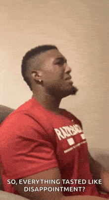 a man in a red shirt is sitting on a couch and making a face .