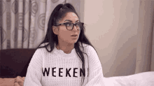 a woman wearing glasses and a sweater with the word weekend on it is sitting on a bed .