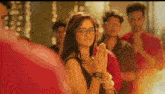 a woman wearing glasses is standing in front of a group of people with her hands folded in prayer .