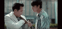 two men are standing next to each other in front of a building and one of them is taking off his jacket .