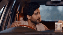a man with a beard is driving a car while holding a cup of coffee .