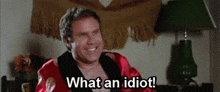a shirtless man in a red robe is sitting in a room and smiling while saying `` what an idiot ! ''