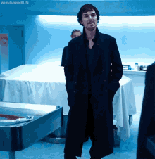 a man in a black coat stands with his hands in his pockets in front of a table with a white cloth on it ..