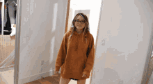 a woman wearing glasses and a brown hoodie is standing in a hallway
