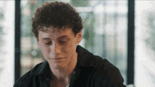 a young man with curly hair is sitting at a table with his eyes closed and a black shirt on .