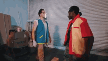 a man in a blue vest is standing next to a man in a red shirt .