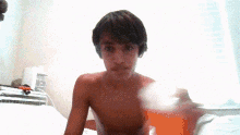 a boy without a shirt is sitting on a bed holding a glass of orange juice