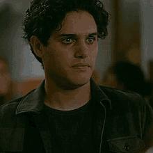 a man with curly hair is wearing a black shirt and a black jacket and looking at the camera .