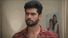 a man with a beard is wearing a red plaid shirt and standing in a room .