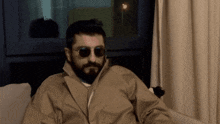 a man with a beard wearing sunglasses and a trench coat sits on a couch