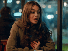 a woman in a green sweatshirt sits at a table with her hand on her chest