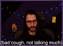 a man with long hair and glasses is sitting in front of a screen that says " bad cough not talking much "