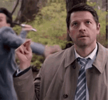 a man in a suit and tie is standing in the woods holding a cigarette .