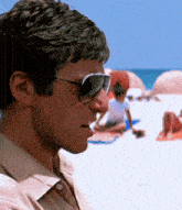 a man wearing sunglasses stands on a beach with people laying on the sand