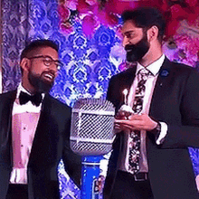 two men in tuxedos are standing next to each other holding a microphone and a candle .