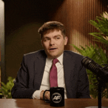a man in a suit and tie is sitting in front of a microphone with a mug that says af on it
