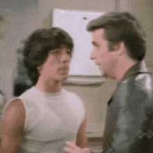 a man in a white tank top talks to another man in a black leather jacket