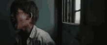 a man in a white shirt is standing in a dark room looking out a window .