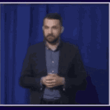 a man in a suit and blue shirt is standing in front of a blue curtain and clapping his hands .