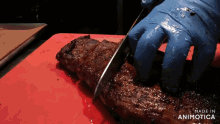a person wearing blue gloves is cutting a piece of meat on a red cutting board that says made in animotica