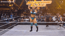 a female wrestler stands in a wrestling ring with her arms in the air in front of a crowd