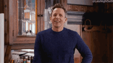 a man in a blue sweater is standing in a kitchen with his hands on his hips and the hashtag late night seth is visible