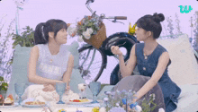 two girls sitting next to each other with a bicycle in the background