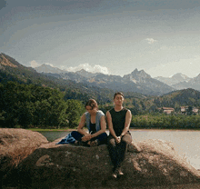 two women sit on a large rock in front of a lake with mountains in the background