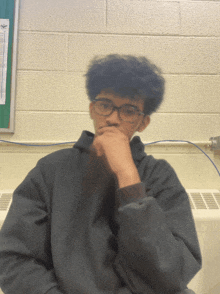 a young man wearing glasses and a hoodie looks at the camera with his hand on his chin