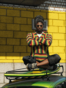 a man in a colorful sweater sits on top of a green car with a surfboard on the roof rack