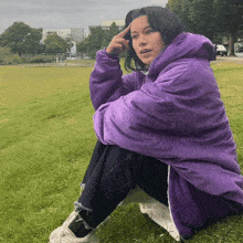 a woman in a purple jacket sits on the grass with her hand on her head