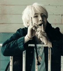 a man with white hair is sitting on a railing talking on a cell phone while holding his finger to his mouth .
