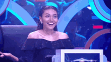 a woman in a black off the shoulder top smiles in front of a blue background