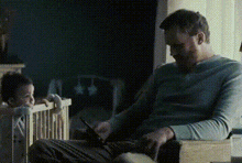a man is sitting in a chair next to a baby in a crib and using a tablet .