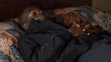 a man in a black jacket is laying on a bed with a patterned blanket