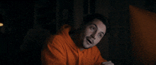 a man in an orange hoodie is sitting in the dark