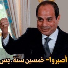 a man in a suit and tie stands in front of a window with arabic writing behind him
