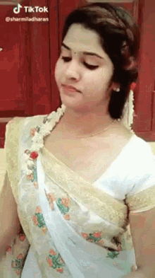 a woman in a white and gold saree is standing in front of a red wall .