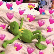 kermit the frog is laying on a bed surrounded by pink hearts and emojis .