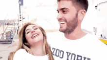 a man and a woman are standing next to each other and smiling . the man is wearing a zoo york shirt .