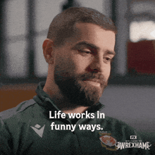 a man with a beard is wearing a green shirt that says " life works in funny ways "