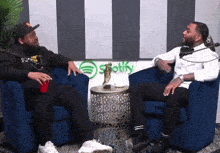 two men sit in blue chairs in front of a spotify sign