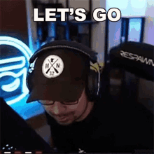 a man wearing headphones and a hat is sitting in front of a computer and says let 's go .