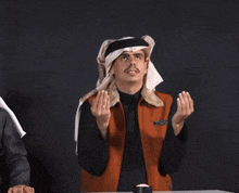 a man wearing a head scarf and a vest is sitting at a table with his hands folded