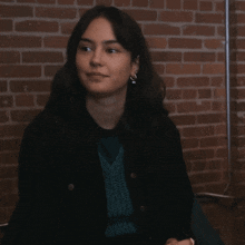 a woman sitting in front of a brick wall wearing a black jacket