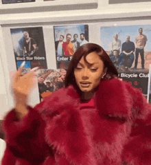 a woman in a red fur coat is standing in front of a wall with posters on it .